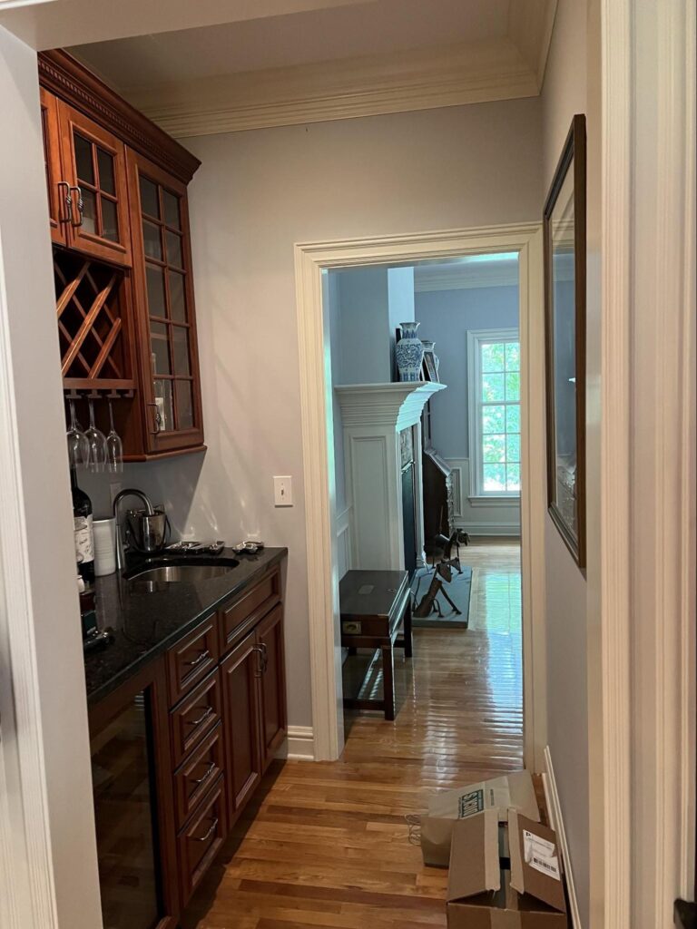 A traditional butler’s pantry with dark wood cabinets, black granite countertop, and built-in wine storage, leading into a formal living room with hardwood floors and classic trim. Cardboard boxes sit on the floor in the hallway, hinting at upcoming renovations.