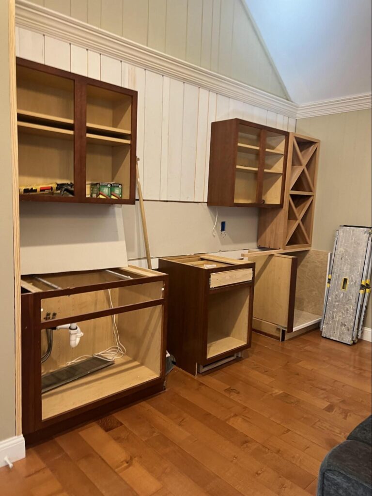 A partially renovated wall with upper and lower cabinets in progress, featuring exposed wood frames and open plumbing below. The warm wood flooring and high, angled ceiling hint at a soon-to-be stylish home bar transformation using repurposed cabinetry.