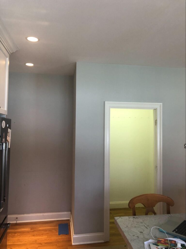 A kitchen corner with light gray walls and wood flooring, showing the space before a coffee station installation. The former pantry space on the right is now an empty recessed nook, freshly painted and brightly lit, awaiting transformation.