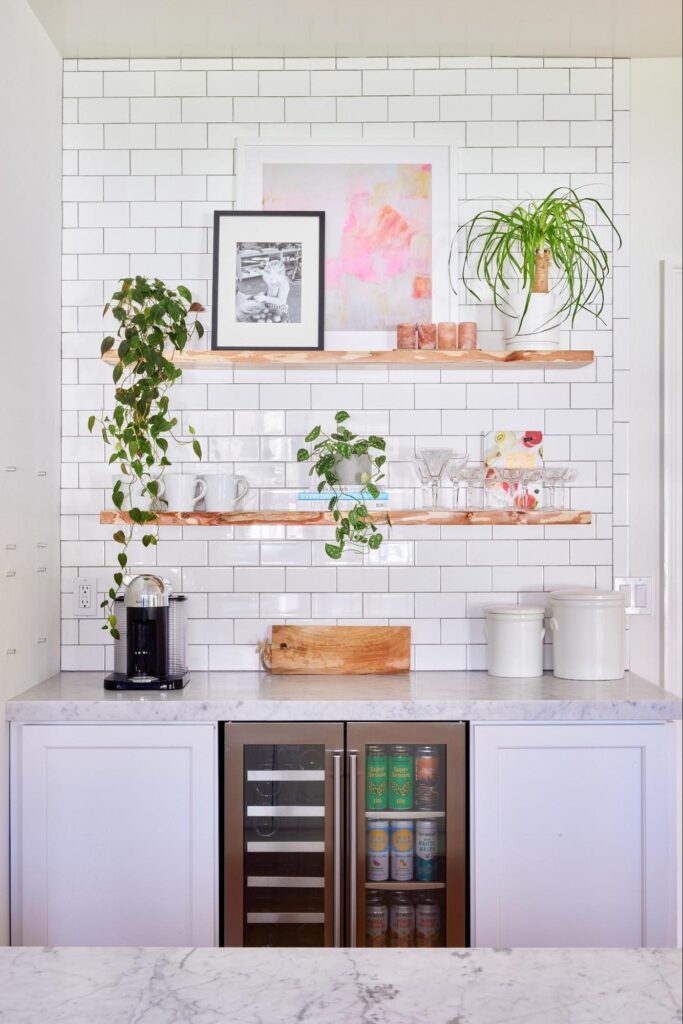 A modern coffee bar featuring white subway tile backsplash, floating natural wood shelves with plants, mugs, and glassware, and a marble countertop with a black coffee machine. Below the counter are dual beverage fridges stocked with drinks, flanked by white cabinetry and ceramic canisters.