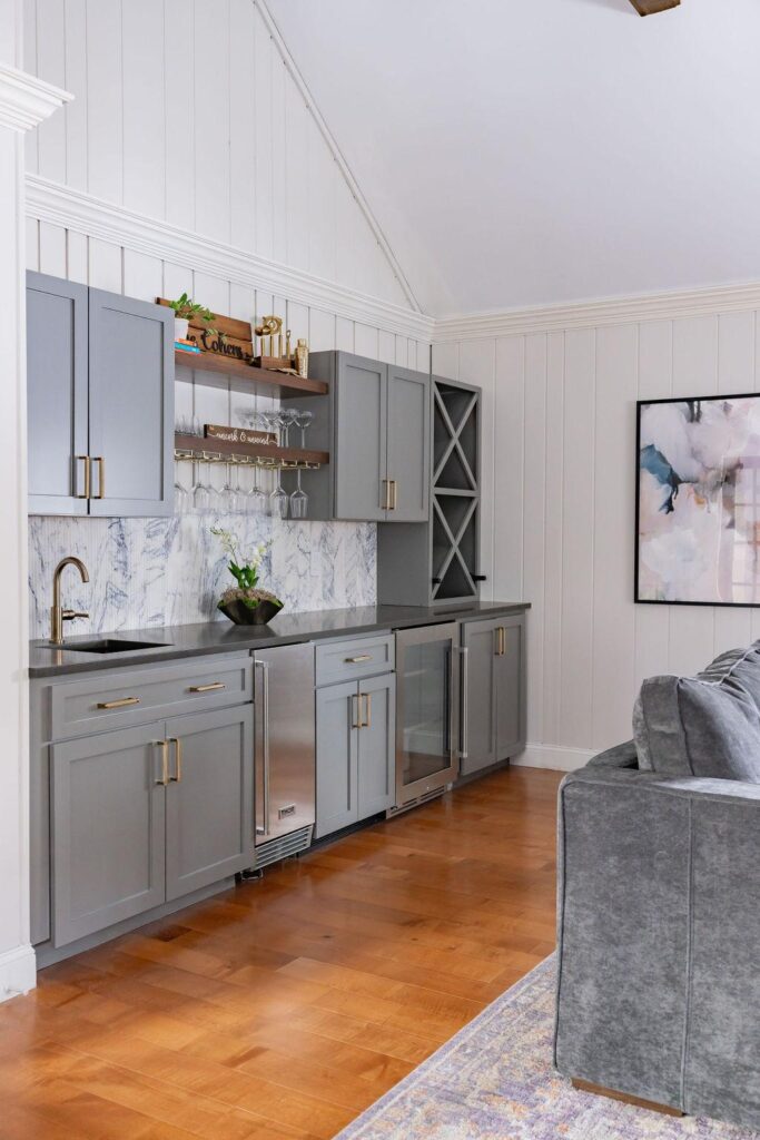 A stylish home bar featuring soft gray cabinets with brass hardware, a fluted marble tile backsplash, and open wood shelves holding glassware and decor. The design includes a sleek black faucet, under-counter beverage fridge, and wine storage, all set against white paneled walls and warm wood flooring.
