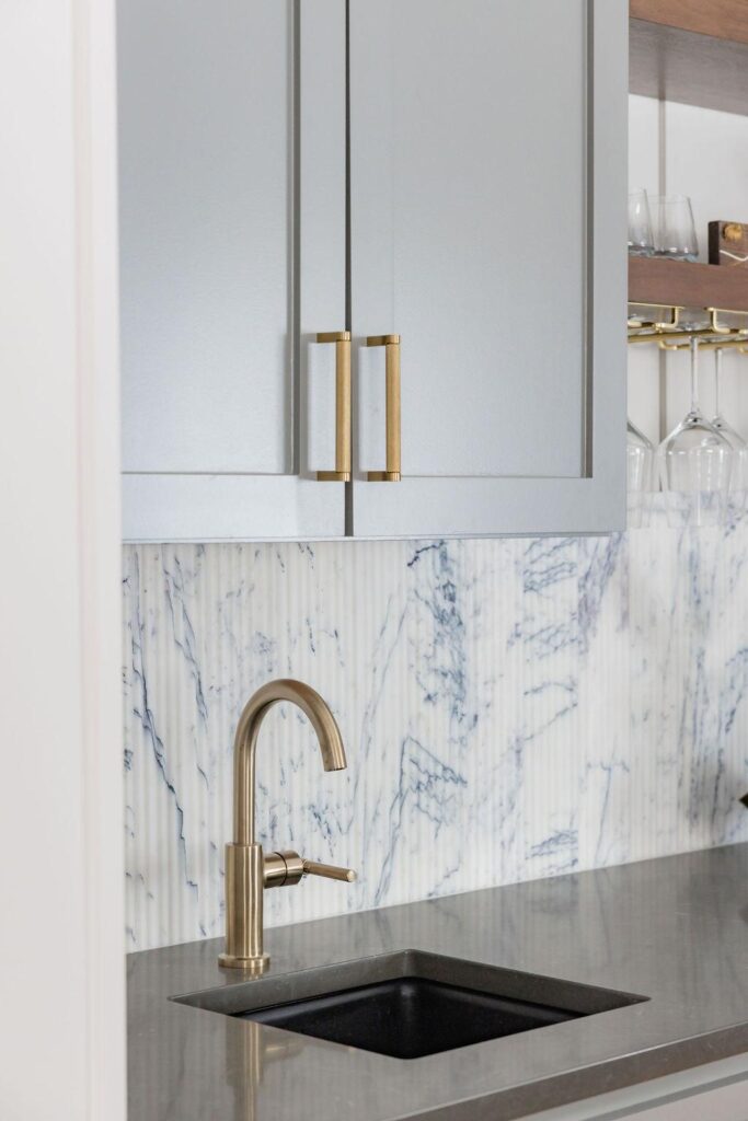 A stylish home bar featuring soft gray cabinets with brass hardware, a fluted marble tile backsplash, and open wood shelves holding glassware and decor. The design includes a sleek black faucet, under-counter beverage fridge, and wine storage, all set against white paneled walls and warm wood flooring.