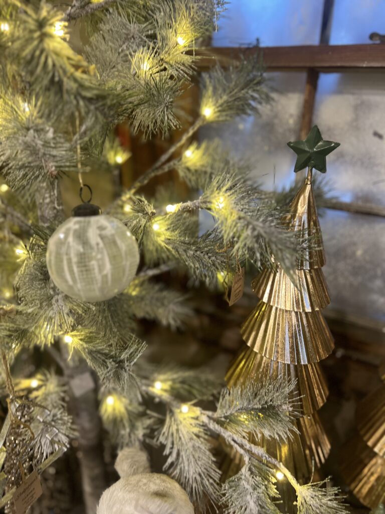 Close-up of a flocked Christmas tree with warm lights, a textured ornament, and a gold sculptural tree decoration beside a frosted window.